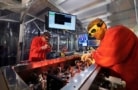 Two men in orange lab coats and laser-safety goggles lean over a bench full of optical components with a screen in the background