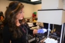 Photo of researcher Nina Vaidya holding an optical concentrator under a machine that is emitting bright, white light