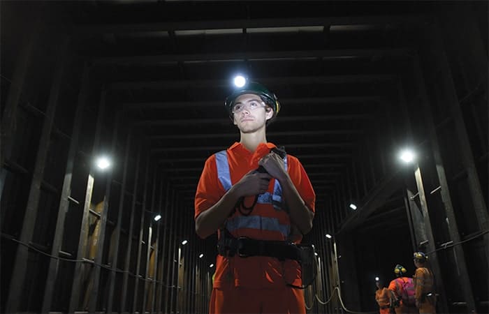 Scientist in underground tunnel wearing bright-orange clothes with fluorescent stripes, a head torch and helmet and mine safety equipment strapped to his waist