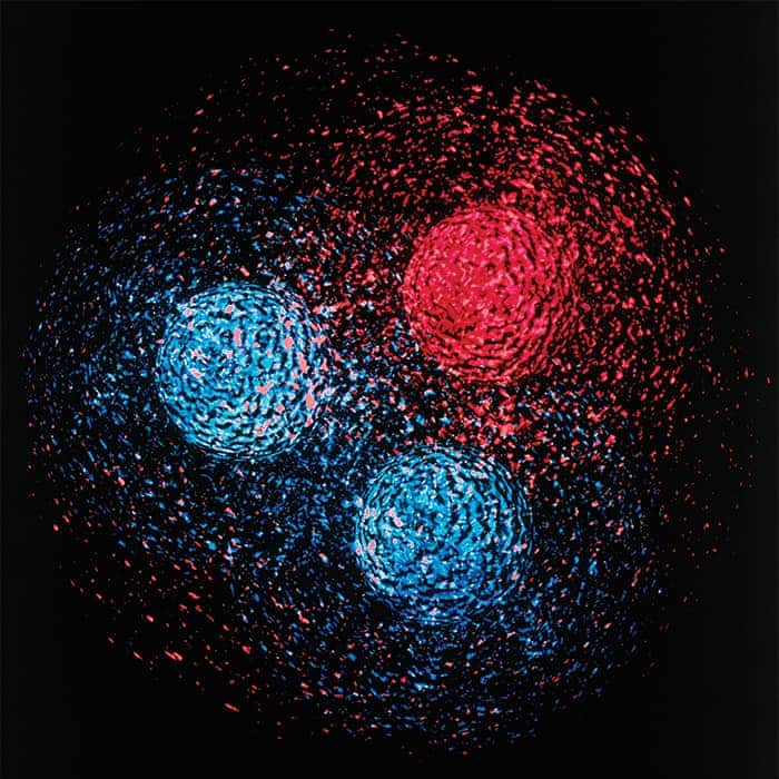 Artwork depicting the proton as a composite particle composed of two fuzzy blue up quarks and one fuzzy red down quark, with bits of red and blue scattered across the background