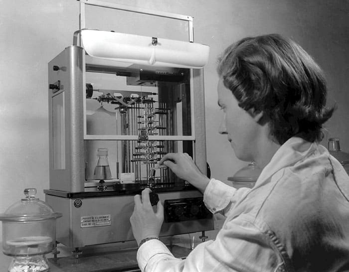 Black-and-white photo of an analytical chemist, Lois Taylor, using a piece of laboratory equipment to carefully transfer material into a small flask